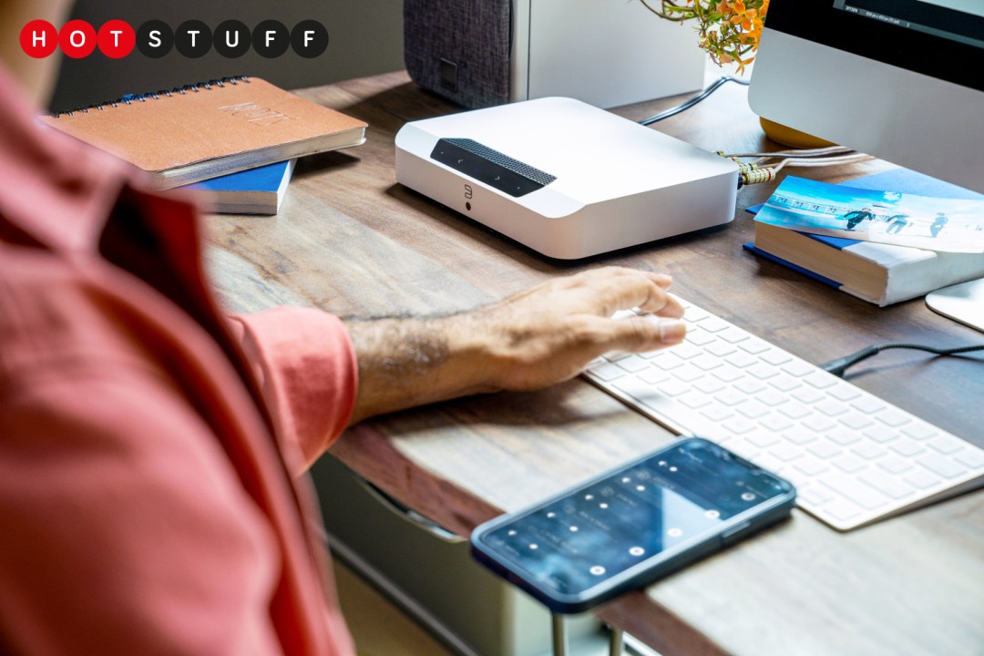 The picture shows a person sitting at a desk, with a smartphone, a computer keyboard, and a white one-box hi-fi.