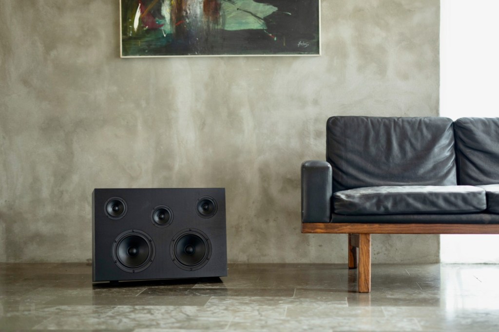 A black, wooden, rectangular Nocs Monolith speaker on the floor next to a sofa. A picture hangs on the wall in the background. 