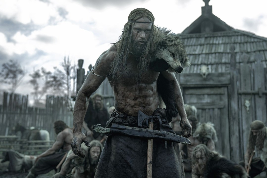 Image shows Alexander Skarsgård in the film The Northman. He's covered in mud and dried blood with a wolf pelt over his shoulder. There is a wooden hut and a group of men behind him.