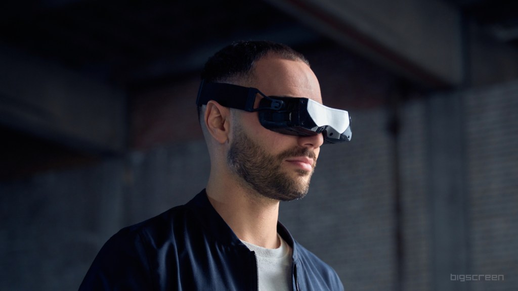 Images shows a man with a beard wearing a Bigscreen Beyond VR headset, a blue jacket and grey t-shirt.