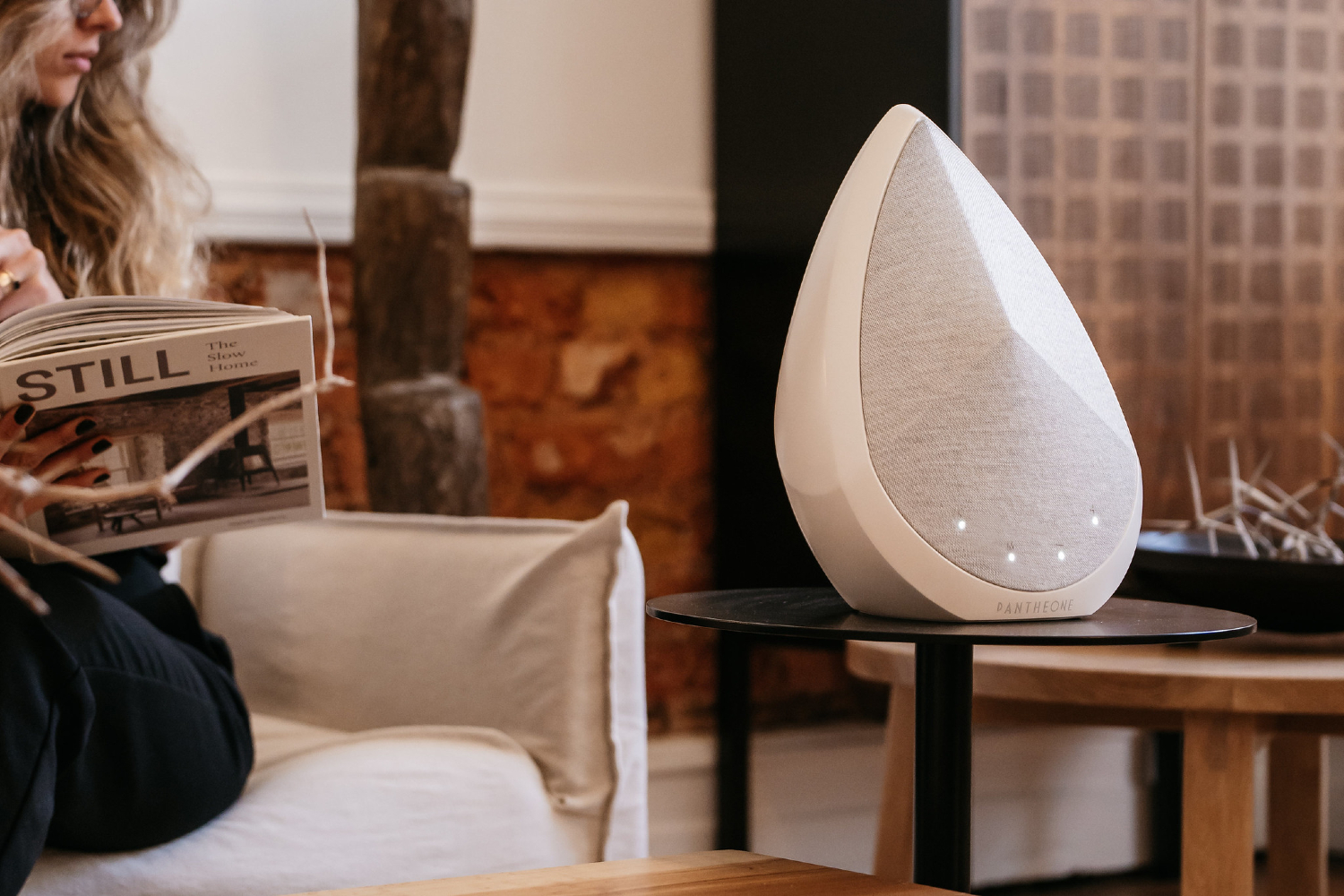 A white Pantheone Audio speaker on a table. In the background, a woman reads a hardback book on sofa.