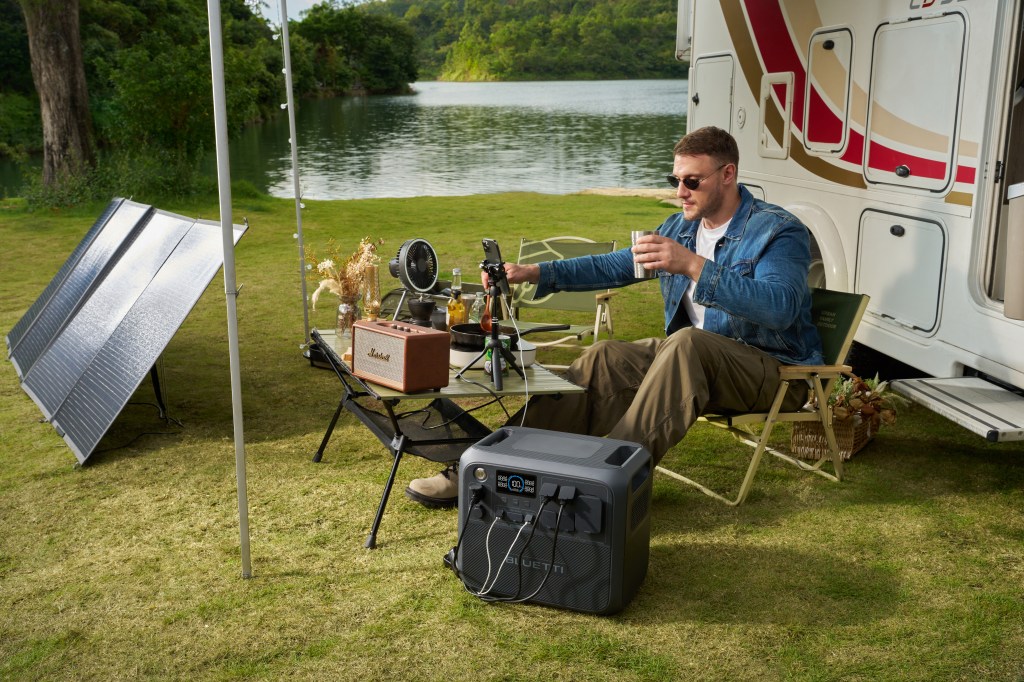 A man camps by a lake with a tableful of gadgets, a solar panel, and a Bluetti power station