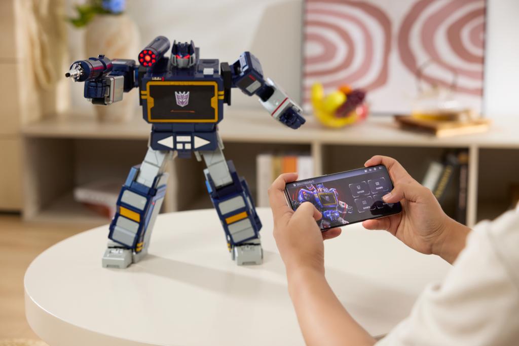 Somebody controlling the Robosen Flagship Soundwave G1 with a mobile phone app. The robot is standing up with one arm stretched out.