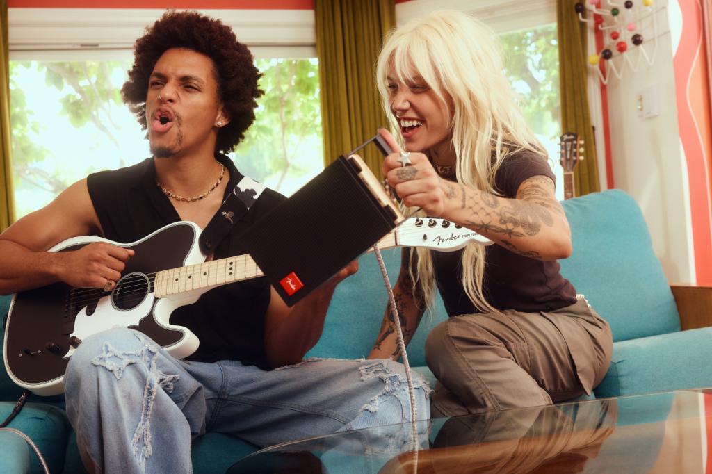 A man and a woman sitting on a sofa. The man is playing a guitar and the woman is holding a Fender ELIE 6 Bluetooth speaker.